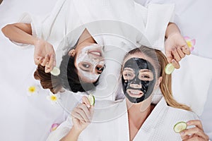 Smiling Caucasian and Asian two girls friends is holding Sliced Ã¢â¬â¹Ã¢â¬â¹cucumber and relaxing with facial masks for skine care